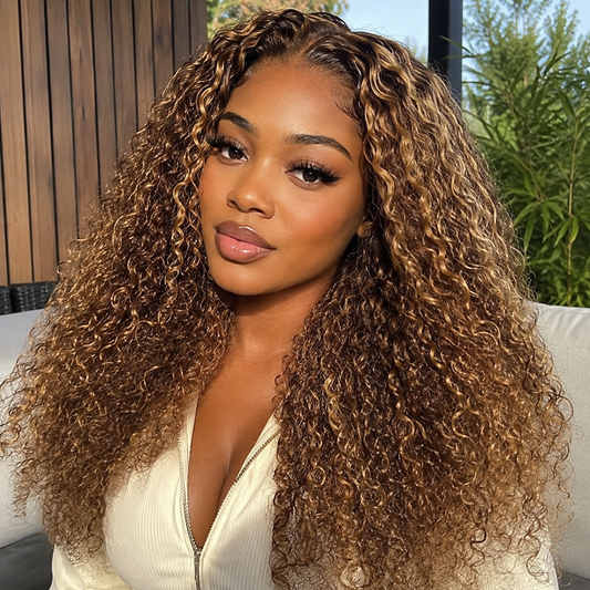 A woman with long, voluminous, tight-curling hair wearing a cream-colored top, photographed in a lifestyle setting with a wooden slatted wall and green plants in the background.